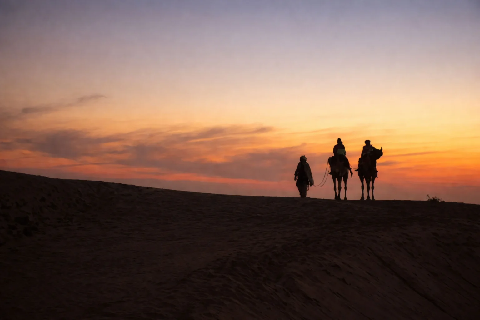 Camels and riders silhouetted on a dune ridge at sunset