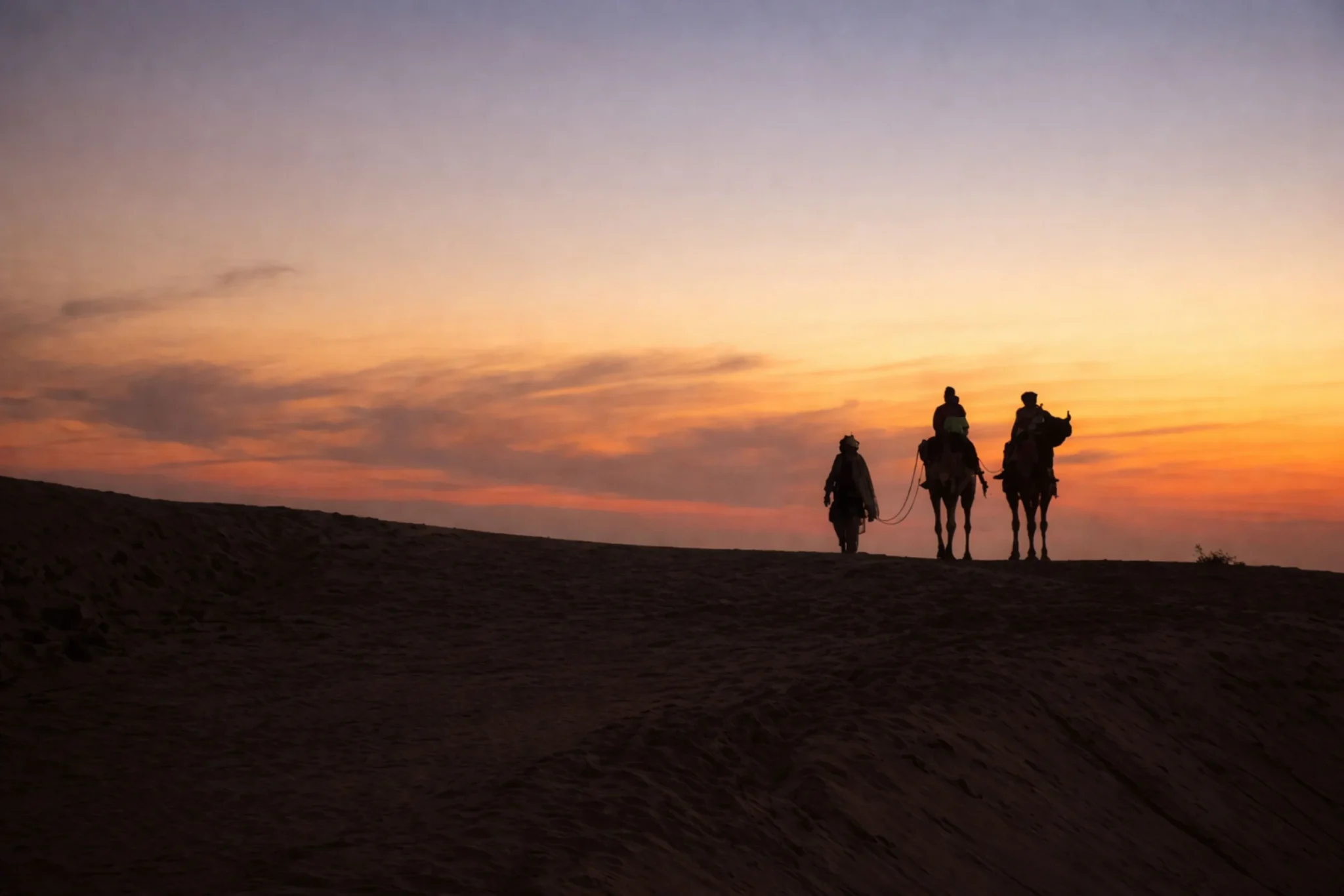 Camels and riders silhouetted on a dune ridge at sunset